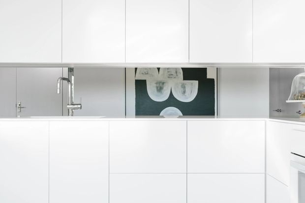 Minimalist white kitchen with extensive flat-panel cabinetry, a white island, integrated appliances, and a stainless steel tap and sink. A dark artwork panel provides contrast behind the sink, while grey mirrored or reflective surfaces appear along the rear wall. The composition is bright, clean, and highly streamlined.