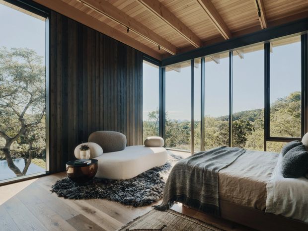 A serene, contemporary bedroom with a low platform bed, textured bedding, a sculptural curved lounge chair, and a small side table. Floor-to-ceiling glazing wraps the room, providing expansive views of a wooded landscape. The room features dark vertical timber wall cladding, light timber flooring, and a warm exposed timber ceiling with beams.
