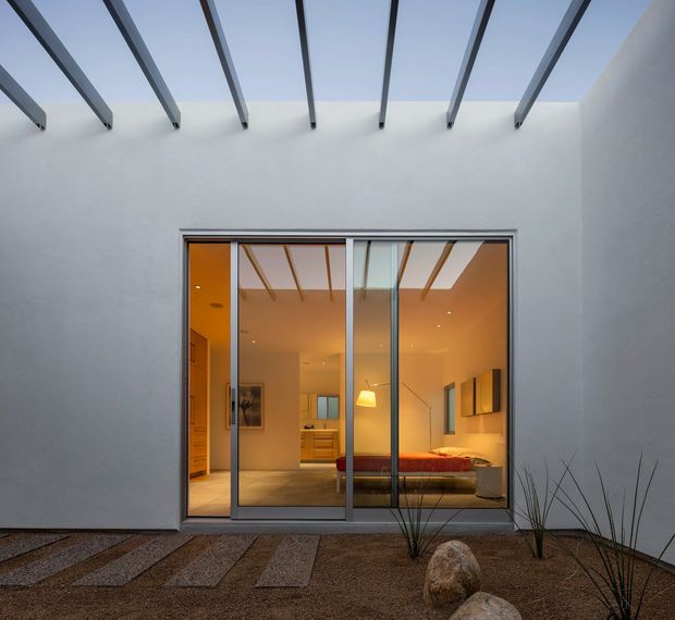 A minimalist courtyard view looking toward a bedroom through large sliding glass doors. White rendered walls enclose the space, with exposed dark roof beams and a partly open skylit roof creating strong linear shadows. The bedroom beyond has warm amber lighting, timber cabinetry, a low bed, and neutral finishes. The courtyard uses gravel, concrete stepping slabs, ornamental grasses, and rocks.