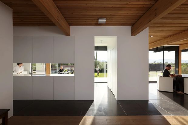 A contemporary open-plan interior with a warm timber-lined ceiling and exposed timber beams. Minimal pale cabinetry forms a kitchen or built-in storage wall on the left, with a narrow horizontal opening revealing people and workspace beyond. Large dark-format floor tiles transition to timber flooring in the foreground. A central doorway leads toward a bright glazed area, while the right side opens to a dining or work area with extensive glazing and views of landscaped grounds.