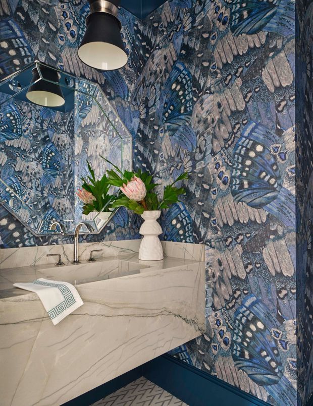 A richly decorated bathroom vanity featuring a large sculptural marble basin and benchtop, polished chrome tapware, a geometric mirror, dark blue cabinetry, and bold blue butterfly-patterned wallcovering. Black pendant lights and a floral arrangement add dramatic decorative accents.