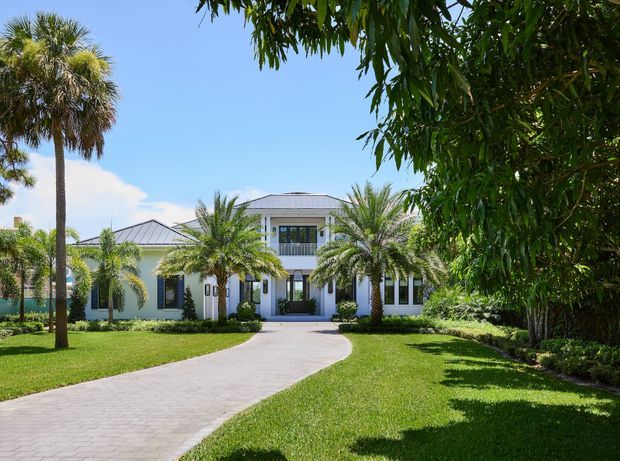 Front exterior view of a large, symmetrical two-storey residence set behind a curved paved driveway. The light-coloured facade features a shallow hipped roof, prominent central entry porch with columns, upper-level balcony, dark-framed windows, and manicured lawn with mature palms and dense tropical landscaping.