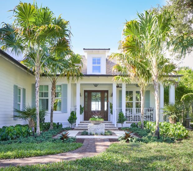 Bright front exterior of a refined, symmetrical two-storey home with a white-painted facade, dark shingle roof accents, a central dormer window, covered entry porch, timber-and-glass front doors, shuttered windows, and lush tropical landscaping with palms. A brick-paved path leads through the garden to the entrance.