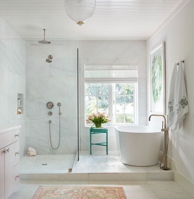 Bright, refined bathroom featuring a glass-enclosed walk-in shower, freestanding white soaking tub, pale marble-look wall and floor surfaces, white cabinetry, a window with a Roman blind, and a white-painted beadboard ceiling. Brass-toned plumbing fixtures and a small teal plant stand provide subtle accents.