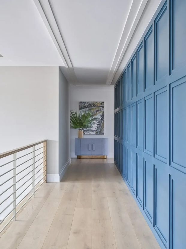 A bright, refined hallway or landing with pale timber flooring, white walls and ceiling, a sleek horizontal metal balustrade, and a dramatic full-height blue decorative wall with raised rectangular panelling and integrated doors. At the far end is a blue-grey console cabinet with a palm arrangement and framed artwork.