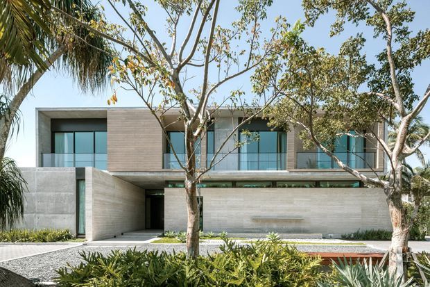 Front exterior view of a contemporary two-storey residence with a flat roof, broad horizontal massing, exposed concrete walls, light timber-look cladding, extensive glazing, and glass balcony balustrades. The home is framed by established tropical landscaping and a paved forecourt.
