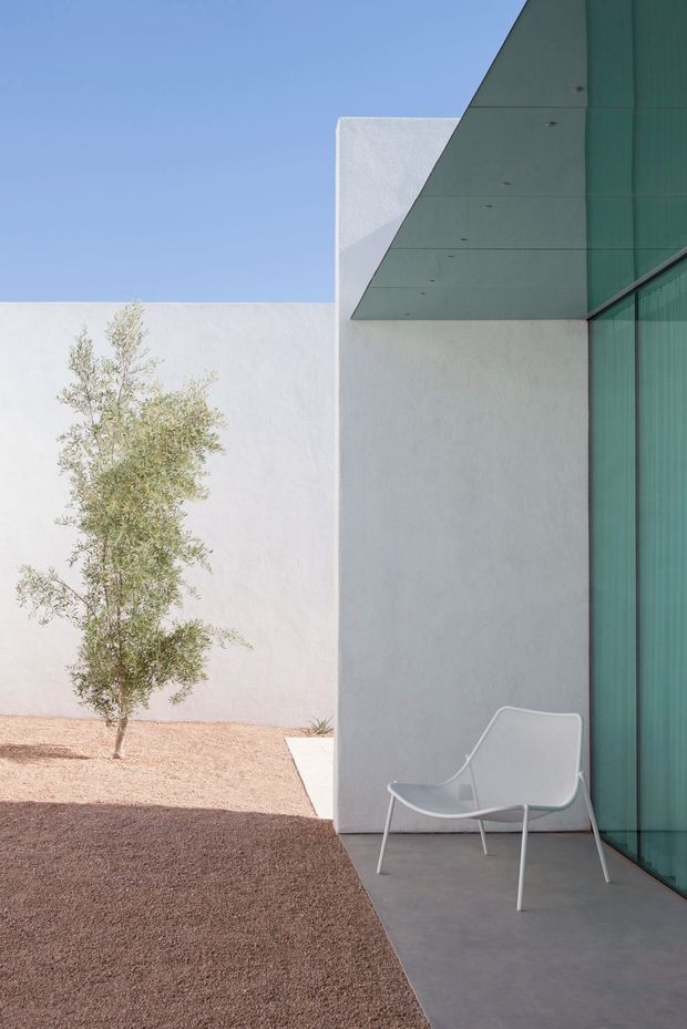 Minimalist modern architectural exterior viewed from a sheltered courtyard or covered outdoor passage. White rendered walls form clean geometric volumes, with a deep projecting green-tinted glazed canopy and full-height glass wall on the right. A simple white lounge chair sits on a smooth grey concrete or paved surface, beside a gravel courtyard with a single slender tree. The composition is calm, sparse, and strongly geometric under a clear blue sky.