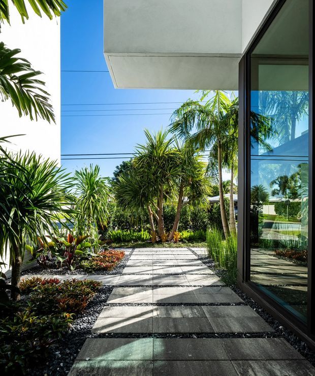 A landscaped exterior walkway beside a modern building, featuring large rectangular grey paving slabs separated by dark gravel, dense tropical planting, and a full-height glazed wall with dark frames. A projecting white concrete roof or canopy and strong sunlight create crisp shadows across the path.