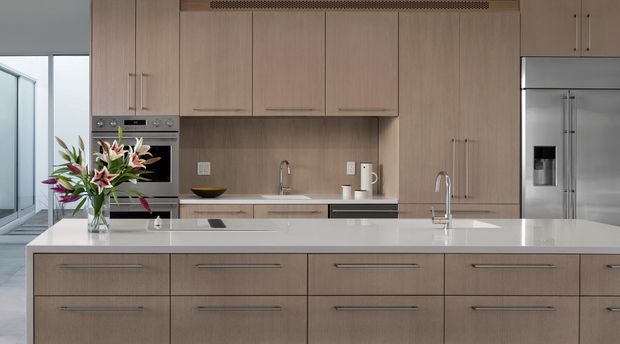 Contemporary kitchen dominated by a large central island with a white stone benchtop and extensive light timber veneer cabinetry. Integrated stainless-steel appliances, twin sinks, minimalist hardware, and a linear ventilation grille create a clean, highly functional fit-out. A glazed area and potted flowers are visible at the side.
