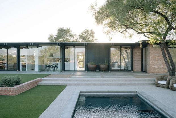 A low, single-storey modern residence viewed from its landscaped courtyard and rectangular swimming pool. The home has extensive floor-to-ceiling glazing in dark metal frames, a flat roofline, pale masonry walls, and a broad paved terrace with steps leading to the building. A dining area is visible through the glazing, while mature trees, clipped lawn, raised brick planting beds, and built-in outdoor seating frame the foreground.