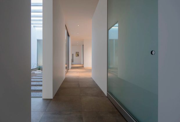 A minimalist interior hallway with a long, narrow circulation path, smooth white walls, recessed ceiling lights, and large-format warm grey floor tiles. Full-height glazing on the right reveals an adjacent room, while an open lightwell or courtyard with horizontal skylight elements is visible on the left. The restrained palette and clean geometry create a calm, gallery-like atmosphere.