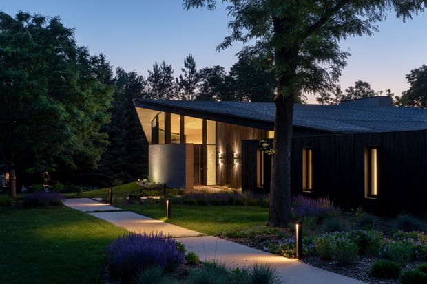 A contemporary single-storey house photographed at dusk, set within a landscaped garden. The dark, low-pitched roof and black vertical exterior cladding contrast with warm light spilling through tall glazed openings. A pale concrete walkway curves through ornamental planting and lawn, with discreet low garden lighting and mature trees framing the facade.