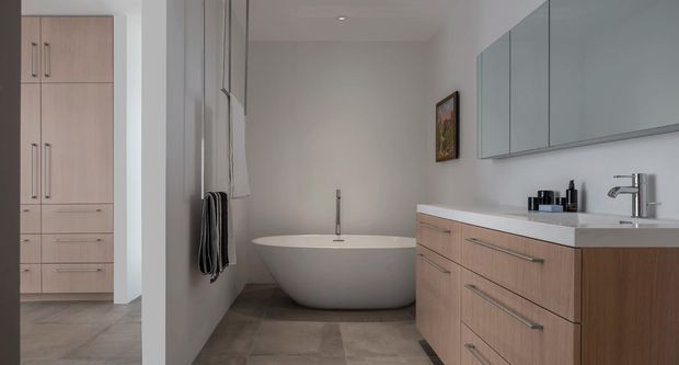 A contemporary bathroom with a freestanding oval bathtub, wall-mounted towel rails, a wide timber-look vanity with integrated drawers, pale blue-grey overhead cabinetry, and a white benchtop with a chrome basin tap. Large-format grey-beige floor tiles and soft neutral walls create a restrained, refined scheme. A built-in storage bank is visible through the opening on the left.