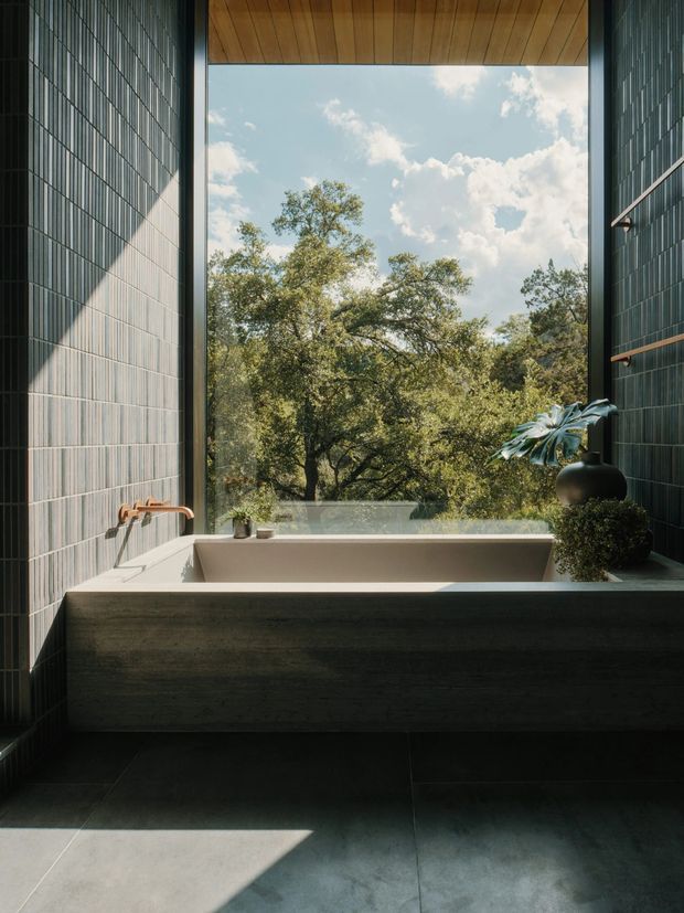 A refined modern bathroom featuring a freestanding rectangular bathtub positioned beneath a large frameless picture window overlooking mature trees. The room has dark vertically ribbed wall tiles, large dark floor tiles, a warm timber-lined ceiling, and a minimalist bronze-toned wall-mounted bath filler. Natural daylight creates strong geometric shadows, with restrained botanical styling on the tub ledge.