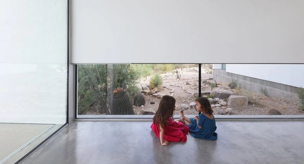 A minimalist interior with a polished grey concrete floor and expansive floor-to-ceiling glazing. Two children sit on the floor beside the window, looking out onto a dry, rocky landscaped garden with desert-style planting. A large white roller blind partially covers the upper window area, creating a quiet connection between the interior and the rugged outdoor landscape.