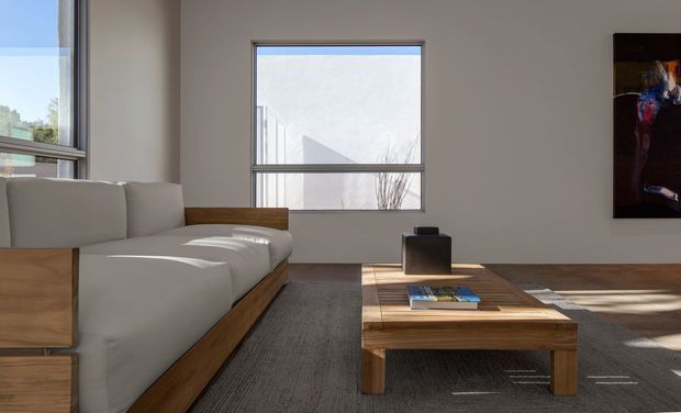 A minimalist living room with a low, pale upholstered sectional sofa set on a timber base, a substantial rectangular timber coffee table, and a dark textured area rug. Large simple windows bring in strong natural light, while a dark abstract artwork hangs on the wall. The room has clean lines, sparse furnishings, and a warm wood-and-neutral palette.