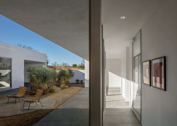 A bright modern courtyard house viewed from an interior circulation space. Full-height glazing opens onto a minimalist xeriscaped courtyard with sculptural desert plants, gravel, concrete paving, and outdoor lounge chairs. The interior corridor has smooth white walls, recessed ceiling lights, large pale floor tiles, framed artwork, and strong linear geometry. The composition emphasizes indoor-outdoor connection and abundant natural light.