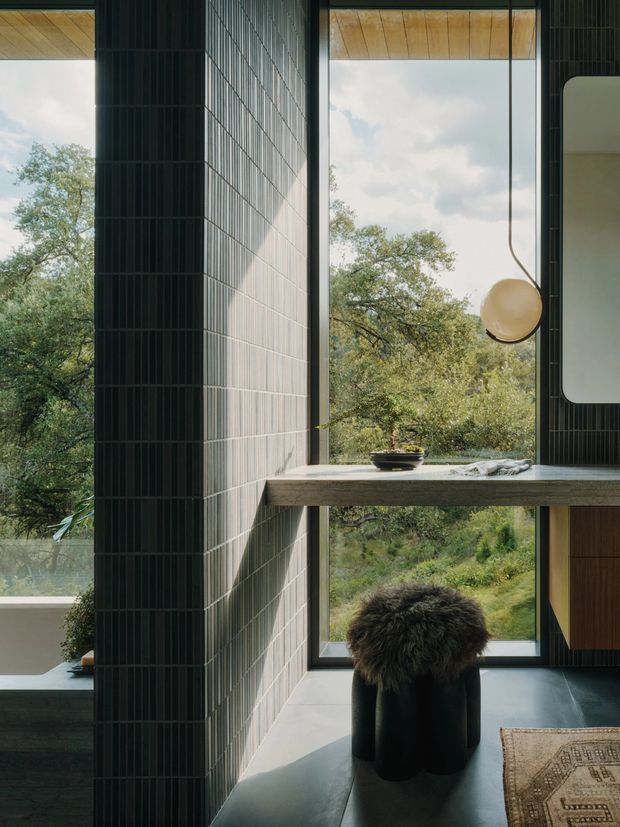 A dark, refined interior corner with floor-to-ceiling glazing overlooking leafy vegetation. Vertical dark wall tiles frame a timber-lined ceiling and a slim stone or concrete worktop/desk projecting across the window. A black sculptural stool with a furry seat, suspended spherical pendant light, integrated dark cabinetry, and a patterned rug create a moody contemporary workspace or living area.