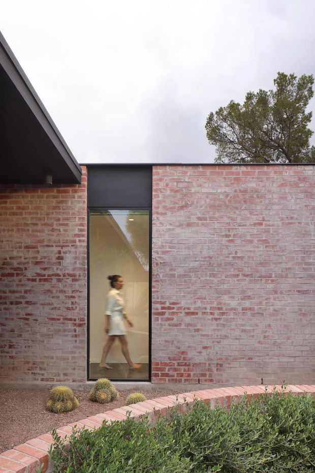 Exterior view of a low, contemporary brick building with a flat roof and a tall, narrow glazed opening framed in dark metal. The pale red and pink brick wall is paired with a dark roof edge and roller blind, gravel landscaping, rounded cacti, clipped shrubs, and a curved brick garden border. A person is visible through the glazing, but the primary subject is the building facade.