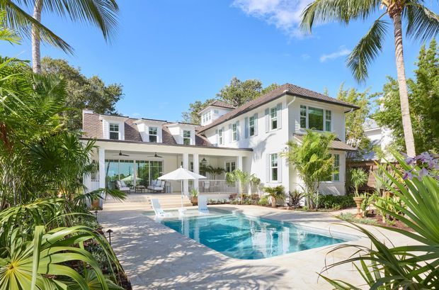 A bright two-storey tropical residence viewed from the landscaped pool area. The white rendered house has a hipped dark tiled roof, multi-pane windows, a covered outdoor entertaining terrace, and a rectangular swimming pool surrounded by palms and lush planting.