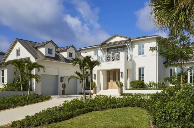 Exterior view of a large white multi-storey residence with multiple gabled roof sections, a covered balcony with decorative railings and columns, attached double garage, and a landscaped front garden with tropical palms. The facade has a bright, refined coastal or traditional character.