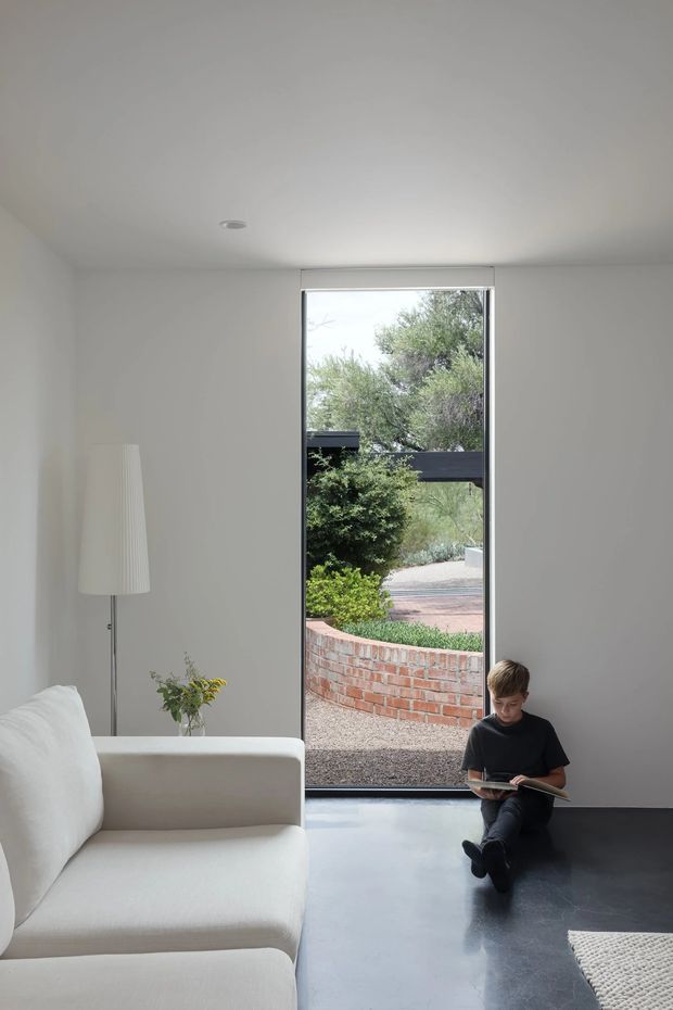 Minimal, light-filled living room with smooth white walls and ceiling, a dark polished floor, and a large full-height window or glazed door opening to a landscaped courtyard. A white sofa, slim floor lamp, and small side table occupy the foreground, while a child sits reading near the glazing. The space has a restrained contemporary architectural character with strong indoor-outdoor connection.