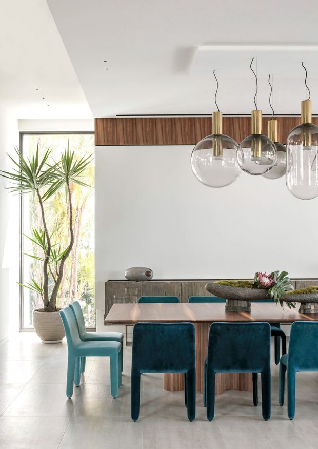 Bright contemporary dining room with a long timber dining table, teal upholstered dining chairs, a timber feature wall and sideboard, sculptural centrepieces, and a cluster of clear glass pendant lights with brass fittings. Full-height glazing beside the room brings in abundant daylight and frames tropical planting outside.