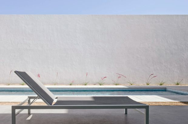 Minimalist outdoor pool area with a sleek dark-framed sun lounger on a pale paved terrace beside a long rectangular swimming pool. A high textured white rendered wall forms the backdrop, with sparse ornamental grasses planted along its base and clear blue sky above. The composition is clean, geometric, and strongly contemporary.
