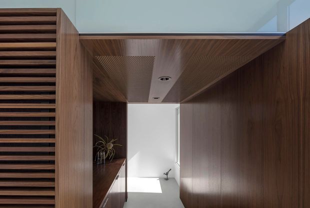 A refined modern interior passage framed by extensive dark timber joinery. Vertical and horizontal slatted timber screens flank the corridor, with integrated cabinetry and a timber-lined ceiling featuring a perforated acoustic panel and recessed downlights. A bright minimalist room is visible beyond, creating strong contrast between the warm wood-lined transition space and the white interior.
