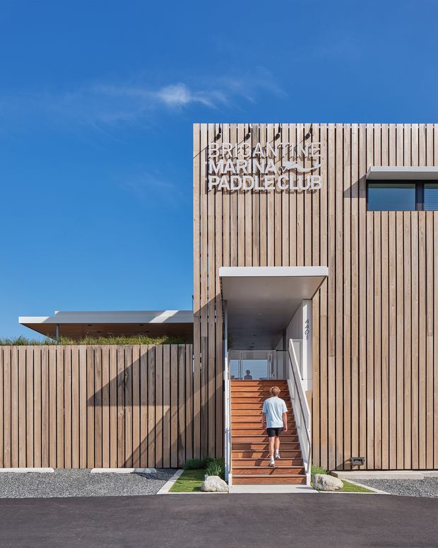 Street-facing exterior of a contemporary marina or paddle club building, featuring a tall facade clad in vertical timber battens, a recessed stair entry, and a low flat-roofed wing behind a timber privacy fence. Clear blue sky and strong sunlight emphasize the clean geometric form. Signage identifies the building as the Bribie Island Marina Paddle Club.