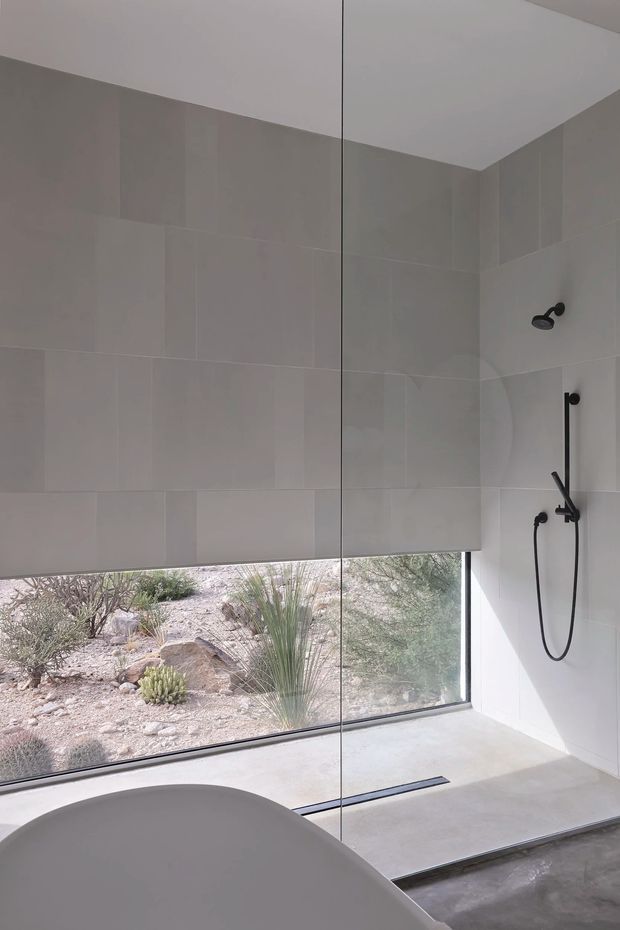 Minimalist contemporary bathroom with a large frameless glass shower enclosure, pale large-format tiled walls, a light concrete shower floor with linear drain, matte black shower fittings, and a freestanding curved bathtub in the foreground. A full-height window overlooks a dry desert-style landscaped setting with rocks and drought-tolerant plants.