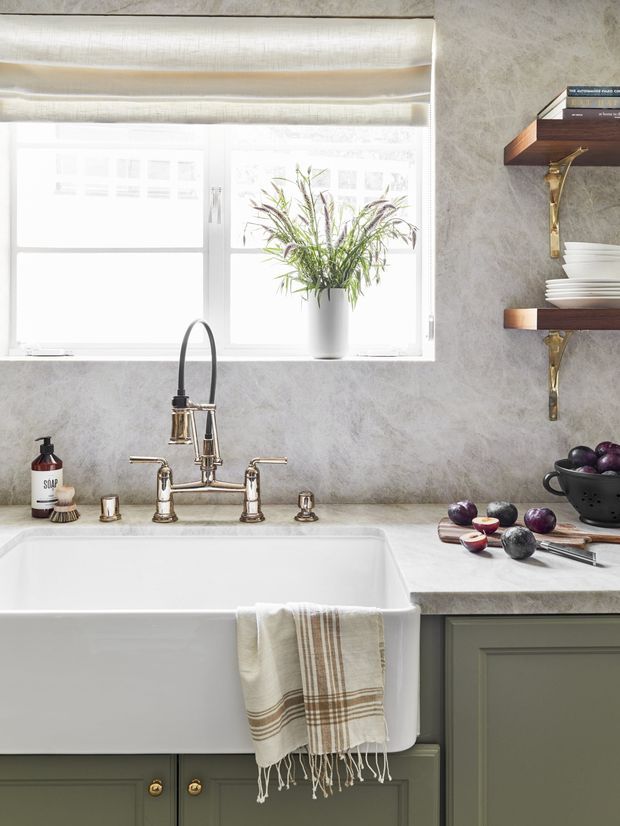 Bright, close-up view of a refined kitchen sink area featuring a deep white farmhouse apron-front sink, polished metal bridge faucet with separate spray fixture, pale marbled stone-look splashback and benchtop, muted green-grey cabinetry with brass knobs, and open timber shelves supported by brass brackets. A window with a light roller blind, potted plant, crockery, fruit and neutral textiles add warmth.