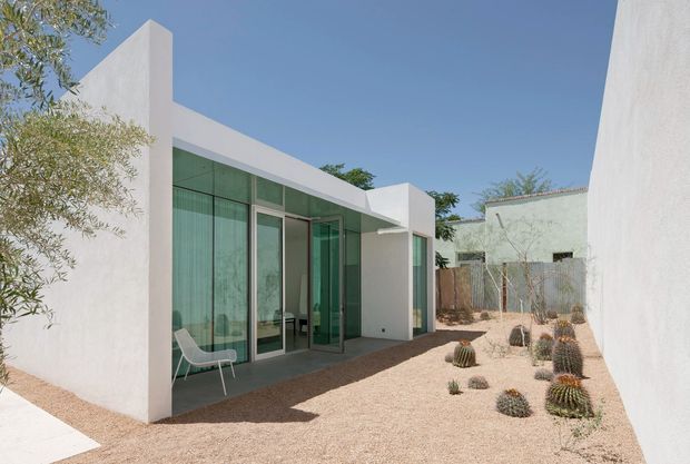Exterior view of a minimalist single-storey residence with crisp white stucco or rendered walls, projecting geometric parapets, extensive green-tinted floor-to-ceiling glazing, and a sheltered terrace. A dry landscaped courtyard with gravel, sculptural cacti, and sparse planting surrounds the building under a clear blue sky.