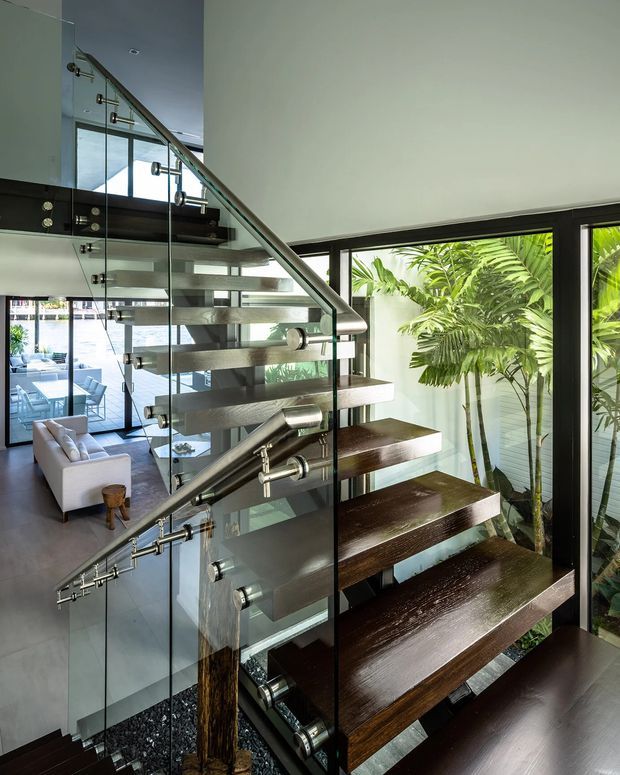 Interior view of a contemporary open-riser staircase with dark timber treads, transparent glass balustrades, and stainless-steel handrails. The stairway is positioned beside large black-framed windows overlooking lush tropical planting, with a modern living area visible beyond.