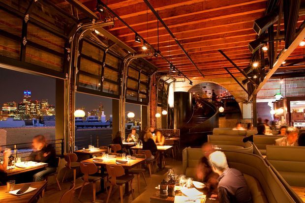 A warmly lit industrial-style restaurant or hospitality interior at night, with exposed timber ceiling beams, dark metal services and hardware, large glazed garage-style doors opening toward a city view, intimate dining tables, upholstered booth seating, and pendant and track lighting. The space has a dramatic, lively atmosphere with a strong indoor-outdoor connection.