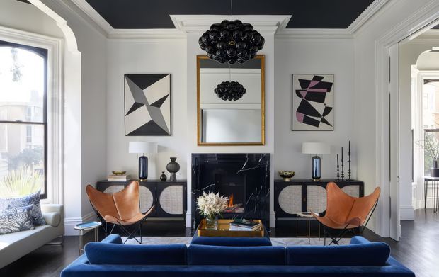 Refined living room with a deep navy ceiling, white architectural mouldings, pale grey walls, a black marble fireplace surround, and a symmetrical arrangement of mid-century-inspired furniture. A large blue sofa faces the fireplace, flanked by cognac leather lounge chairs and dark sideboards, with a sculptural black pendant, geometric artwork, and a gold-framed mirror adding visual interest.