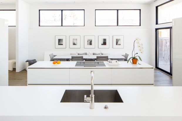Bright open-plan kitchen viewed from behind a large white island with an integrated stainless-steel sink and tap. The space features streamlined white cabinetry, a dark cooktop, pale timber flooring, black-framed horizontal windows, full-height glazed doors, a dining area, framed artwork, and a small indoor plant. The room has a clean, minimalist contemporary appearance with abundant natural light.