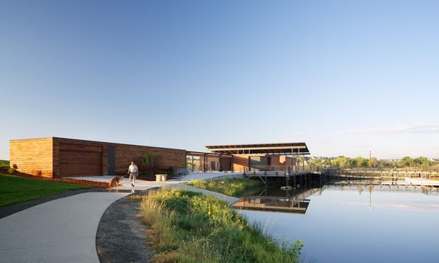 Wide exterior view of a low, contemporary timber-clad building beside a calm reflective body of water. The single-storey structure has long horizontal glazing, a projecting flat or shallow roof canopy, dark timber wall cladding, and a paved pedestrian path leading toward the entrance. Landscaped grasses and lawn frame the waterfront setting.