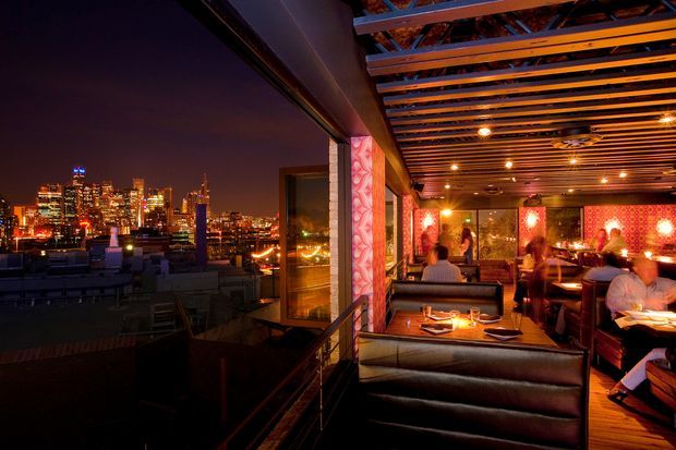 Night-time view of a stylish rooftop dining venue, with illuminated city skyline visible through expansive glazing on the left and a warmly lit restaurant interior on the right. The dining area features upholstered booth seating, tables, patterned red-orange wall panels, exposed dark timber ceiling battens, and dramatic ambient lighting.