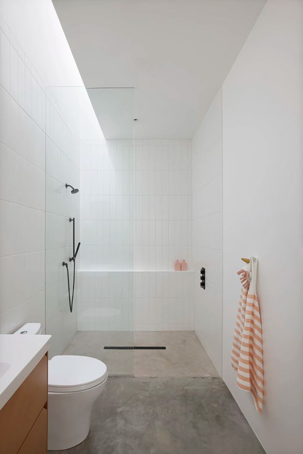 A bright, minimalist bathroom with a walk-in shower occupying the rear of the room. White tiled walls, a pale concrete-look floor, black shower fittings and drain, a wall-mounted towel hook, and a compact vanity with toilet are visible. A long skylight or clerestory opening provides natural light above the shower.