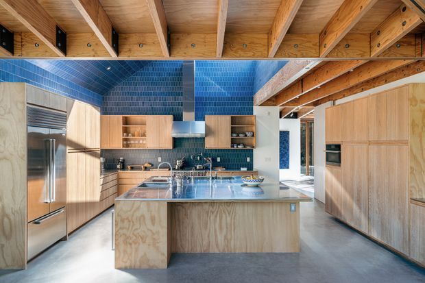 A spacious contemporary kitchen with a large central island, extensive pale timber cabinetry, stainless-steel appliances, and a vivid blue tiled splashback and wall finish. The exposed timber beam ceiling and plywood joinery create a warm, natural contrast with the cool blue tiles and polished grey floor.