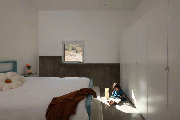 A contemporary bedroom with a low platform bed, white bedding, and a brown throw blanket. The room has white walls and full-height white built-in wardrobe cabinetry, a dark wood-toned wall panel behind the bed, a small turquoise bedside table, and a compact high-set window. A child is reading on the floor in a sunlit corner, with minimal decor and strong natural light.