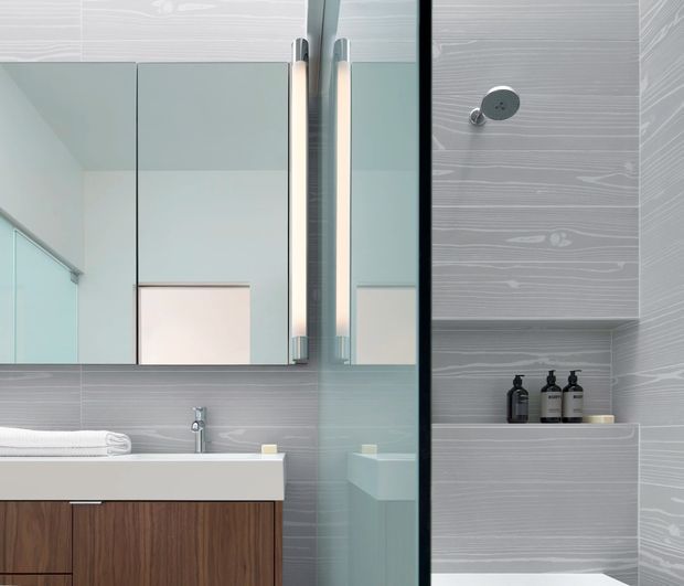 Contemporary bathroom vanity and shower area with a floating wood-front cabinet, white benchtop basin, mirrored wall cabinet, integrated vertical lighting, and a frameless glass shower enclosure. The shower walls feature pale grey horizontally textured tiles, creating a clean, cool-toned finish.