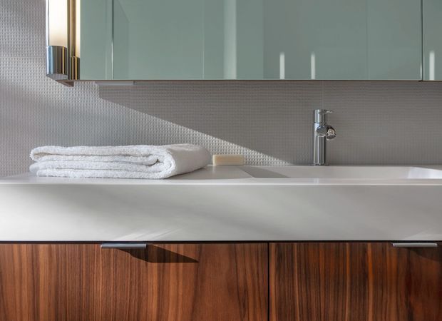 Close-up view of a refined bathroom vanity with a white integrated basin, chrome mixer tap, warm dark-wood cabinetry, pale blue-grey overhead cabinets, and a small textured mosaic tile splashback. Folded white towels rest on the countertop. The composition is clean and contemporary with strong horizontal lines and soft natural light.