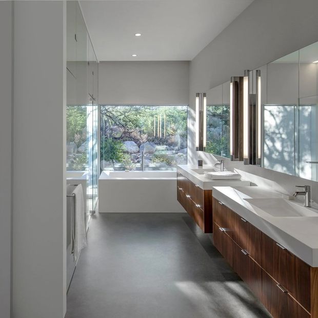 A spacious contemporary bathroom with a freestanding white bathtub positioned beside full-height glazing overlooking a landscaped rocky garden. A long double vanity features white stone countertops, integrated basins, warm dark timber cabinetry, large wall mirrors, and vertical illuminated mirrors. The room has smooth grey flooring and walls, recessed ceiling lights, and abundant natural light.