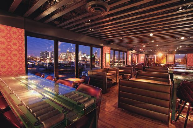 A moody, upscale restaurant or bar interior with extensive floor-to-ceiling glazing overlooking a brightly lit city skyline at night. The space features rows of dark leather booth seating, a long bar counter with stools, warm timber flooring, exposed dark timber ceiling joists, dramatic warm pendant and recessed lighting, and bold patterned red-orange wall panels and artwork.