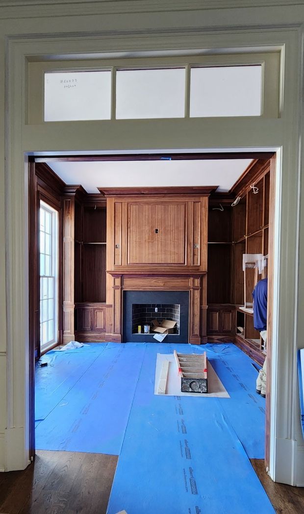 View through a wide doorway into a formal room under renovation. The room features extensive dark timber wall panelling and built-in cabinetry surrounding a central fireplace with a black brick-lined firebox and a large paneled timber overmantel. Tall glazed doors or windows appear on the left, while blue protective floor covering, exposed work materials, and partially completed joinery indicate construction or restoration work. The surrounding doorway has painted trim and a row of clerestory windows above.