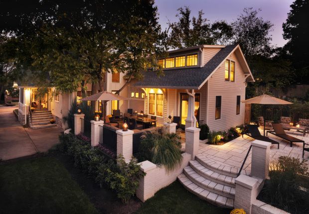 Twilight exterior view of a substantial multi-level residential home with a steep gabled roof, light painted weatherboard cladding, dark shingle-like roofing, and warm illuminated windows. The landscaped property includes broad paved terraces, curved steps, low brick garden walls and columns, outdoor dining and lounge areas, and umbrellas. Mature trees frame the house and create an inviting evening atmosphere.