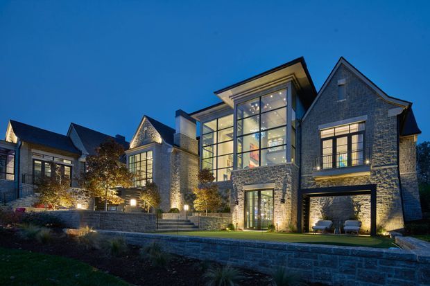 Dusk exterior view of a large multi-wing residential home with prominent grey stone masonry, steep gabled roof forms, tall black-framed glazed sections, and warm architectural lighting. The landscaped foreground includes tiered stone retaining walls and planted areas. A covered opening at ground level appears to be a garage or sheltered outdoor area.