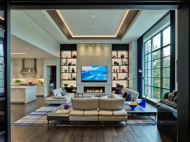 A refined open-plan living room viewed from a dark-framed glazed opening, featuring a large sectional sofa facing a wall-mounted television and linear fireplace. Built-in open shelving flanks the fireplace, while a light-toned kitchen with island is visible to the left. Tall black-framed windows bring in abundant natural light, and the room has dark timber flooring, a layered area rug, and a recessed ceiling with timber trim and concealed LED lighting.